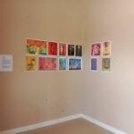 The Techspressionism show in a gallery room in Khiva, Uzbekistan in a beige-walled corner displays colorful abstract artworks arranged in two rows, with a text description on the left wall and twelve vibrant prints showcased on the right.