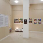 The Techspressionism show in a gallery room in Khiva, Uzbekistan features beige walls adorned with text panels and colorful photographs. A white bench sits near the wall, while a tall column stands centrally under the bright, even lighting.