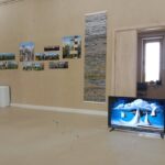 The Techspressionism show in a gallery room in Khiva, Uzbekistan features photos on a beige wall, a tall textured artwork beside an open door, and a TV on the floor displaying white sailboats on blue water beneath a cloudy sky.