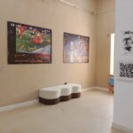 The Techspressionism show in a gallery room in Khiva, Uzbekistan with beige walls, two colorful abstract posters, a white and brown bench, a window with blinds, plus a wall-mounted QR code and monochrome artwork.