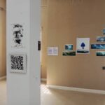 The Techspressionism show in a gallery room in Khiva, Uzbekistan displays various artworks on beige walls, including a black and white portrait, a large poster, landscape photos, and a visible QR code on a pillar. Sunlight streams in from a door on the right.