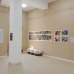The Techspressionism show in a gallery room in Khiva, Uzbekistan with beige walls, featuring two white benches, large text panels, and a series of colorful photographs and artworks. The room is well-lit and minimalistic.