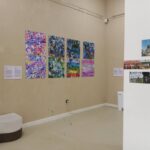 The Techspressionism show in a gallery room in Khiva, Uzbekistan displays eight colorful abstract paintings in two rows. To the right is a white pillar with four small photos and to the left is a modern white bench. Two text panels are mounted beside the art.
