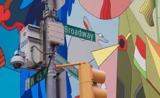 A street sign at the corner of Broadway and West 45th Street in New York City, next to a traffic light and security camera, with a colorful mural in the background.