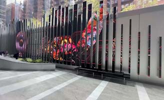 A modern outdoor plaza features a tall metal fence with vertical slats and colorful abstract artwork visible through the gaps, surrounded by buildings and greenery.