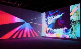 A dark room with two intersecting walls displaying colorful, abstract digital projections featuring vivid streaks of red, pink, blue, and other bright tones creating a dynamic, immersive visual effect.