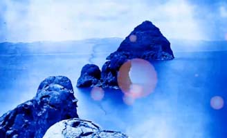 Rocky islands rise from bright blue water under a hazy sky, with sunlight creating lens flare effects on the image. The distant shoreline is faintly visible in the background.