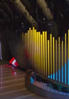 A stage with tall, colorful vertical light bars in yellow and blue, set against a dark background, with a few people seated at the front.