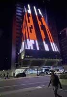 A tall building at night displays bright, geometric red, white, and black light patterns on its facade; several people and cars are visible on the street in front.